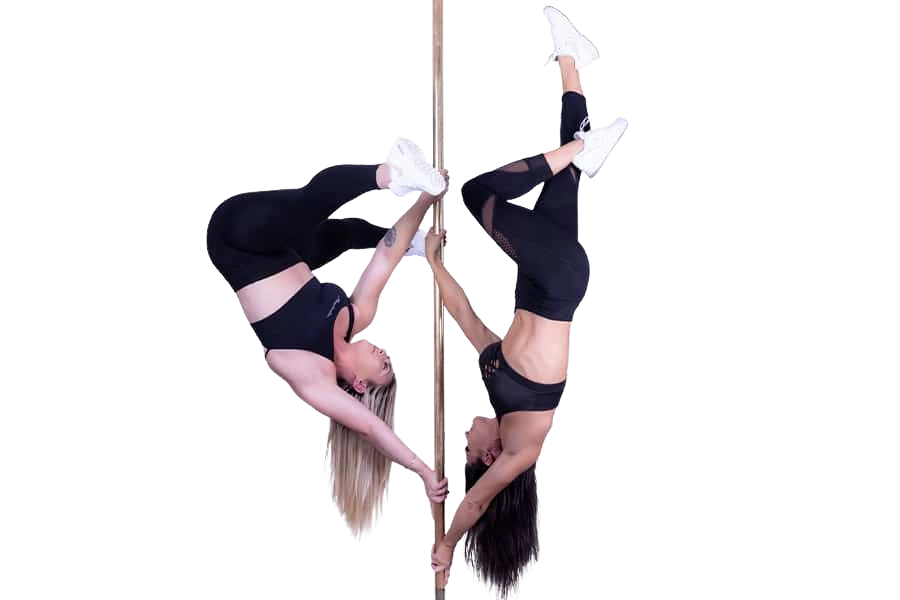 Two women performing inverted pole fitness poses, hanging upside down on a vertical pole while wearing black athletic outfits and white sneakers against a black background.